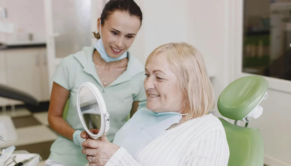 Elderly female dental looks at teeth in a hand-held mirror while dentist watches